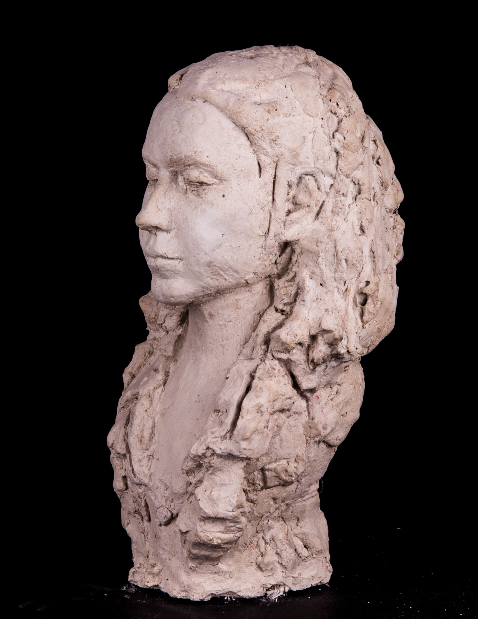 Gipsowy portret kobiety z profilu na czarnym tle, ukazujący detale faktury i niedokończone fragmenty rzeźby.