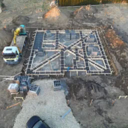 Z lotu ptaka: wykop pod fundamenty domu z drewnianym szalunkiem i zbrojeniem, obok ciężarówka z minikoparką, palety bloczków betonowych i sterta ziemi.