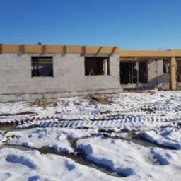 Wznoszenie ścian parterowego domu z bloczków betonowych w zimowej scenerii; widoczne otwory okienne i zbrojenie wieńca na fundamencie.