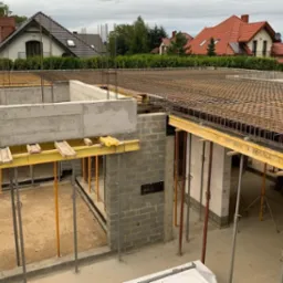 Budowa domu w trakcie: betonowe fundamenty, ściany z bloczków, zbrojenie stropu widoczne z góry, podpory szalunkowe, w tle domy jednorodzinne.