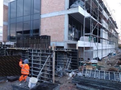 Pracownik w pomarańczowym kombinezonie przy ustawianiu szalunków na budowie domu z dużymi oknami, widoczne rusztowania i materiały budowlane.