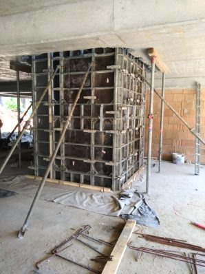 Szare szalunki kolumny w trakcie betonowania, wsparte na stemplach budowlanych, widoczne zbrojenia i elementy konstrukcyjne w surowym stanie.