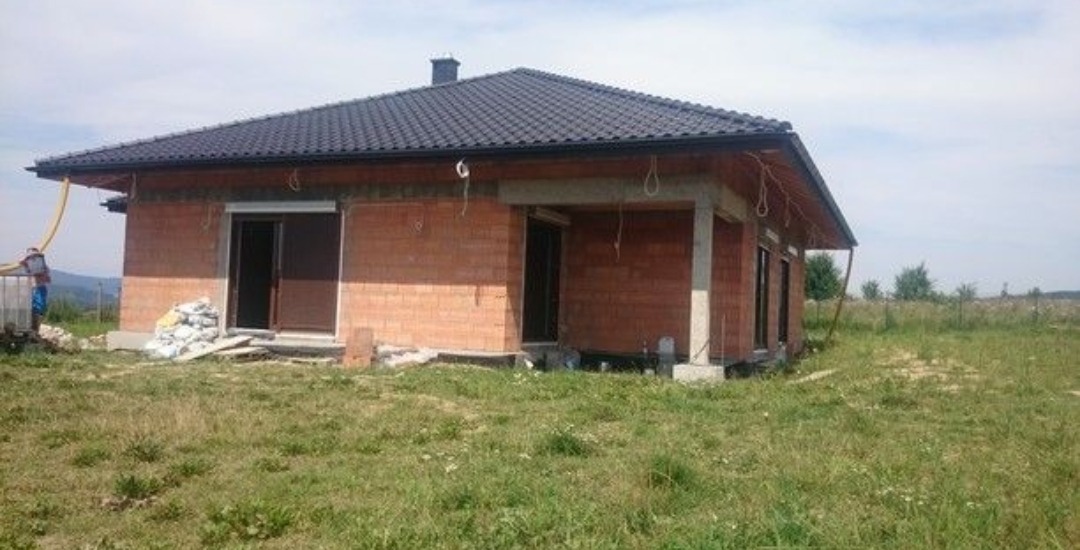 Surowy, jednokondygnacyjny dom w trakcie budowy z czerwonej cegły, z ciemną dachówką, widoczne worki z zaprawą i rura doprowadzająca materiał, otoczony wysoką trawą.
