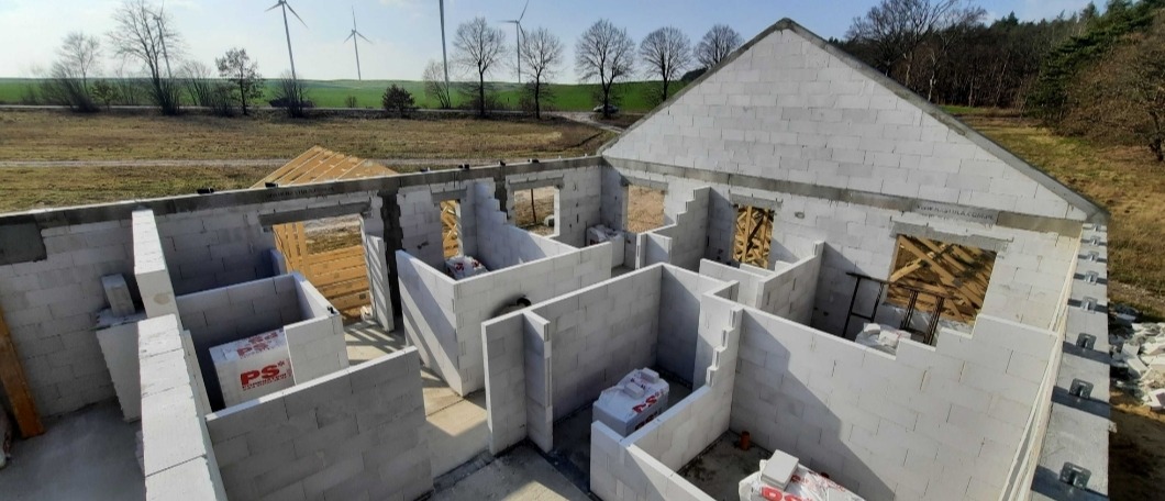 Wnętrze domu w trakcie budowy, ściany wymurowane z białych bloczków, widoczne pakiety materiałów budowlanych w pomieszczeniach, w tle krajobraz z turbinami wiatrowymi.