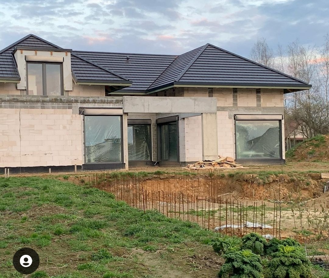 Surowy stan domu jednorodzinnego z ciemną dachówką, widoczne bloczki konstrukcyjne, duże okna zabezpieczone folią i wykop pod fundamenty z wystającymi prętami zbrojeniowymi.