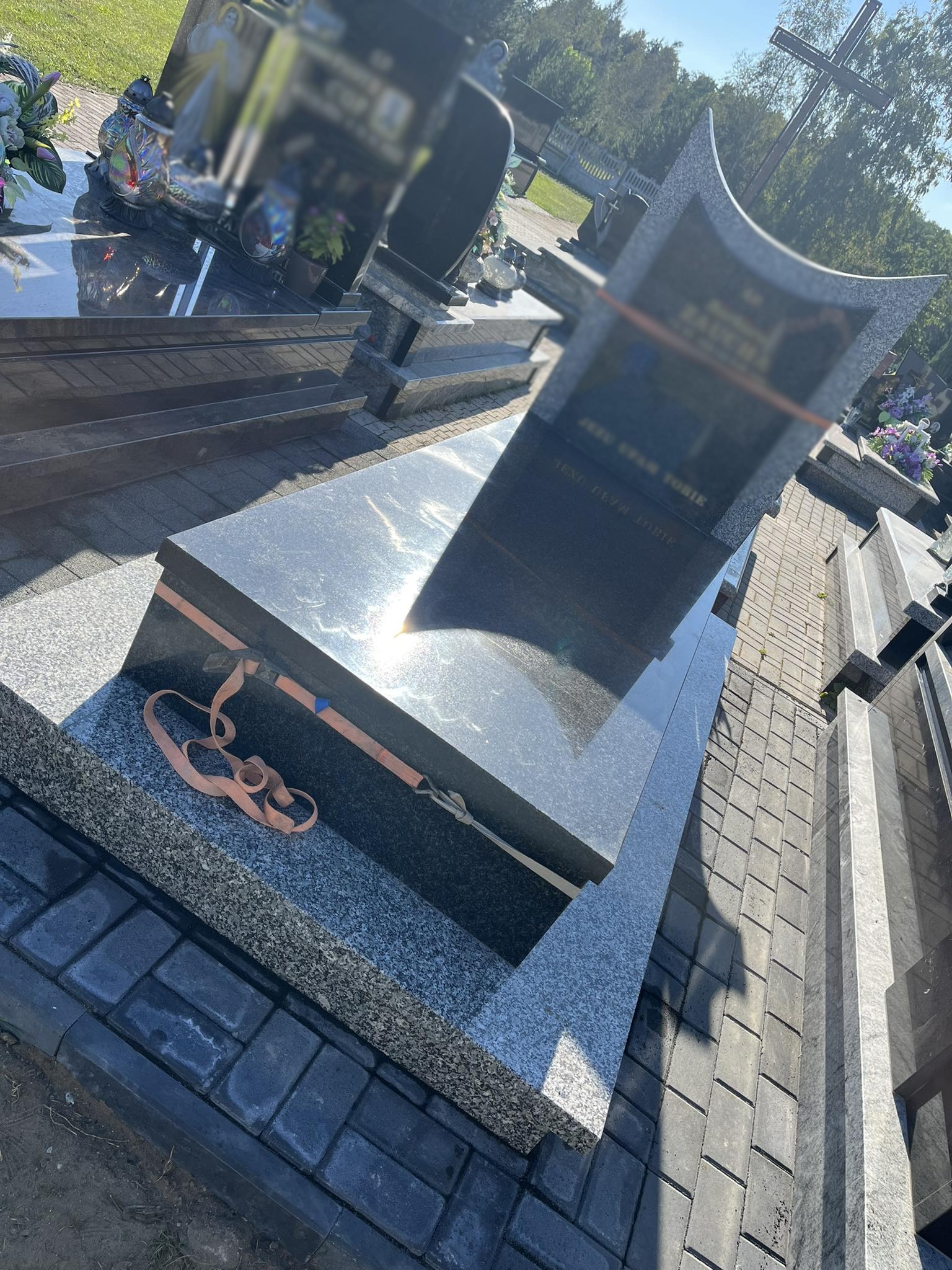 Czarny, błyszczący pomnik na cmentarzu, zabezpieczony pasem transportowym. Widoczne inne nagrobki i krzyż w tle.