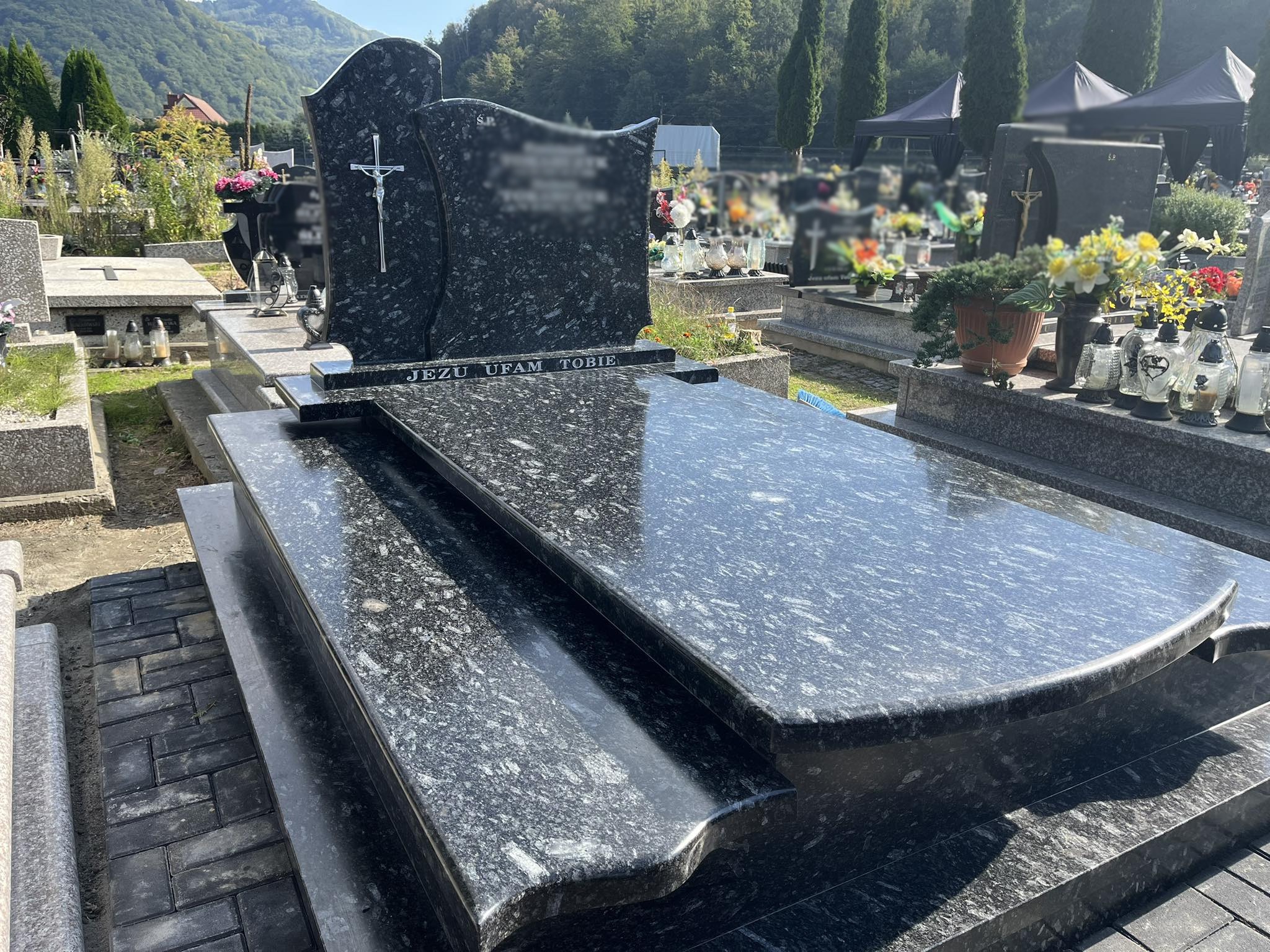 Czarny, granitowy pomnik z napisem 'Jezu ufam Tobie' i srebrnym krucyfiksem, ustawiony na cmentarzu z widokiem na góry w tle.