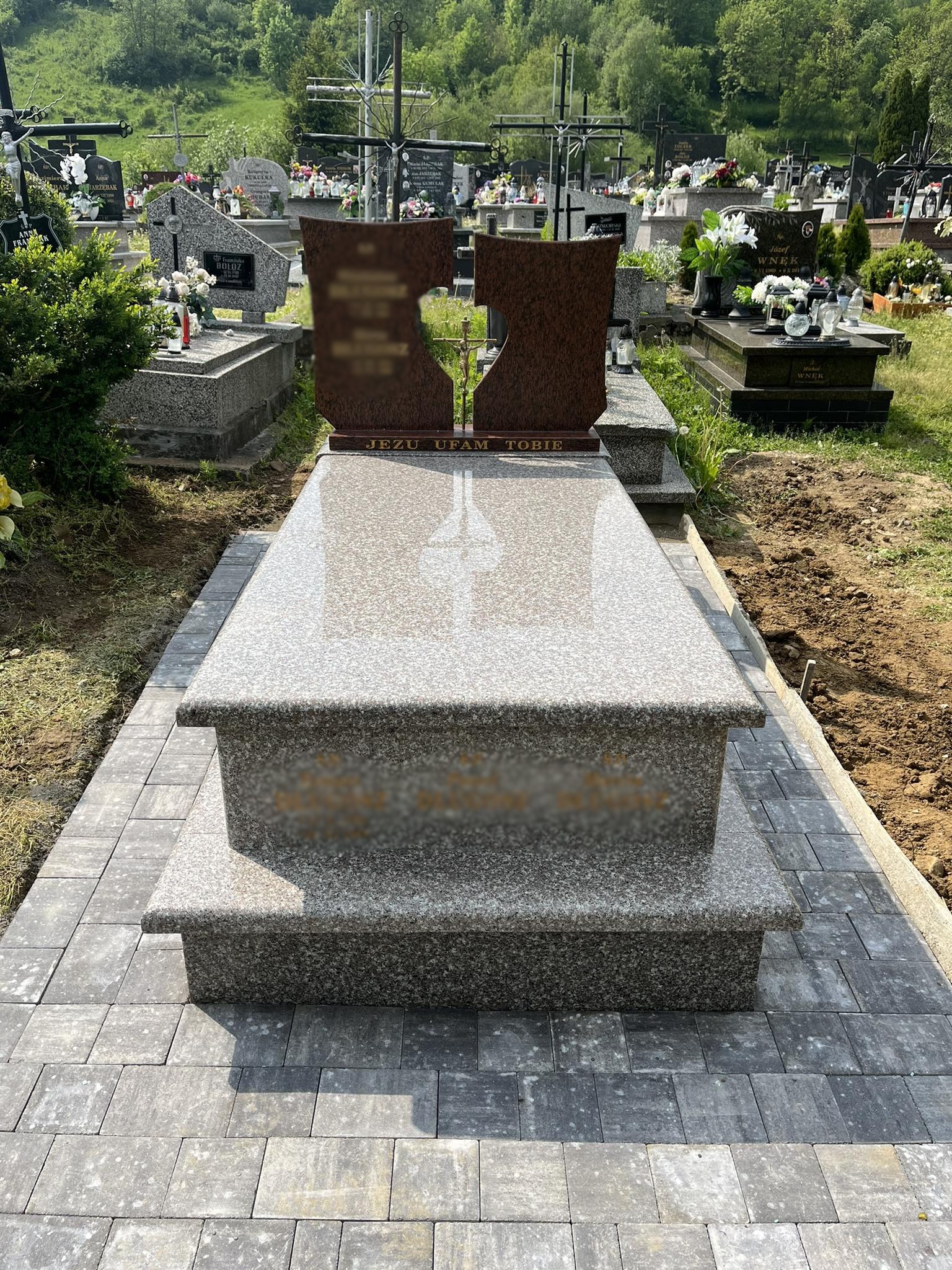 Jasnoszary, granitowy pomnik z brązowymi elementami w kształcie serca, otoczony świeżo ułożoną kostką brukową na cmentarzu w słoneczny dzień.