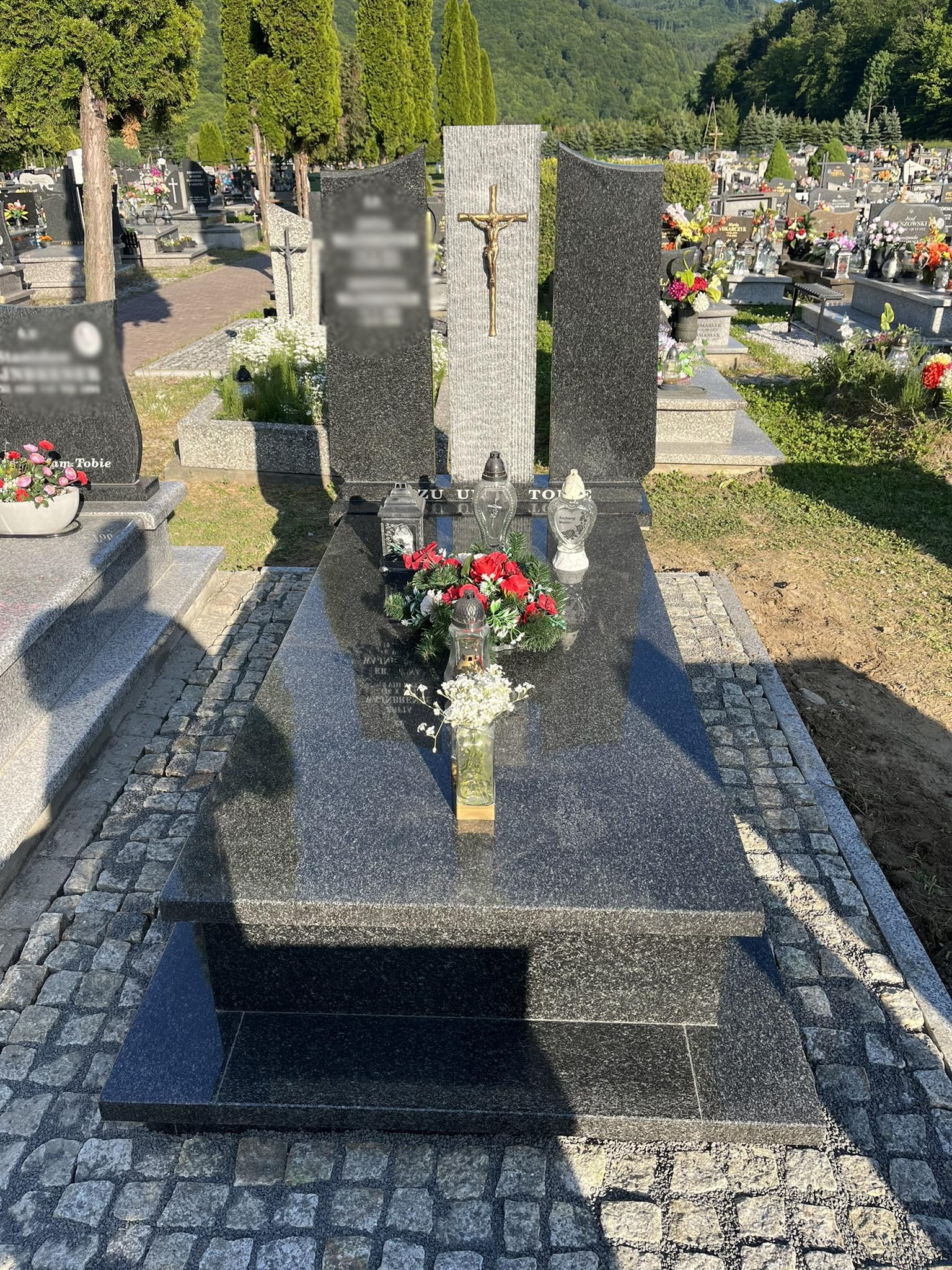 Czarny, granitowy nagrobek z krzyżem, otoczony kostką brukową, z kwiatami i zniczami, na tle cmentarza i zielonych wzgórz.