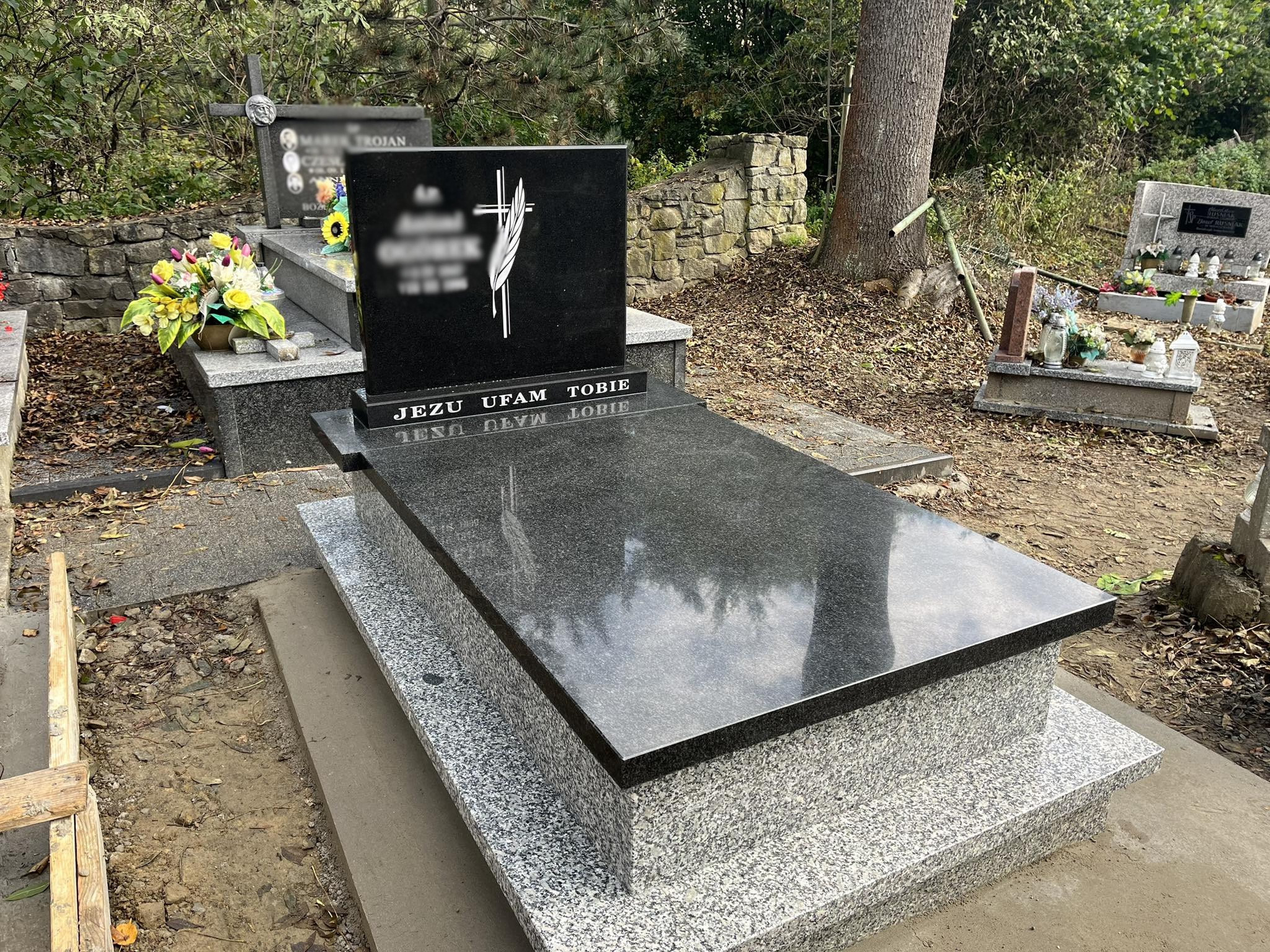 Nowy, granitowy pomnik z napisem 'Jezu ufam Tobie' na cmentarzu, widoczne kwiaty i inne nagrobki w tle.