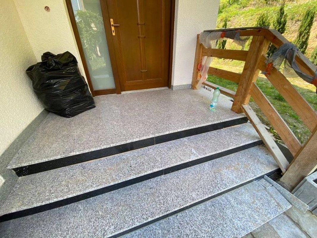 Granitowe schody wejściowe z czarnymi akcentami, widoczny czarny worek na śmieci przy ścianie i prowizoryczna drewniana barierka po prawej stronie podczas montażu.
