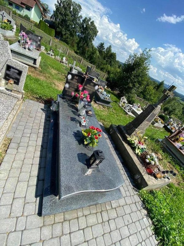 Granitowy nagrobek z kwiatami i lampionem na cmentarzu, otoczony kostką brukową i zielenią, widok z góry pod kątem.
