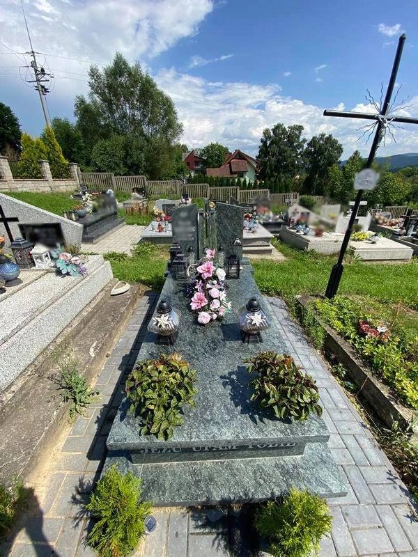 Szary, granitowy pomnik na cmentarzu, z kwiatami i lampionami, otoczony kostką brukową i zielenią, w tle inne nagrobki i drzewa.