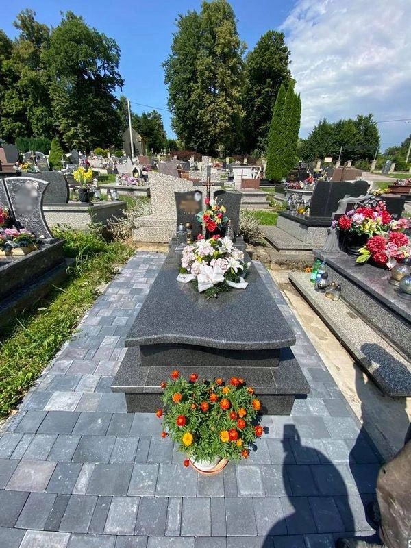 Granitowy pomnik z dwupoziomową podstawą, ozdobiony kwiatami i wieńcem, otoczony kostką brukową na cmentarzu w słoneczny dzień.