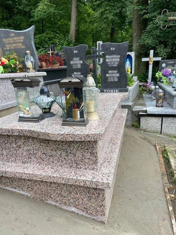 Granitowy pomnik z trzema stopniami, otoczony świeżo wylanym betonem i lampionami, na tle drzew i innych nagrobków.