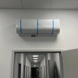 Klimat_pro Petro Fediuk - Nowo zamontowana klimatyzacja Haier, jeszcze z folią ochronną i taśmą, w biurowym korytarzu z widoczną osobą w odblaskowej kamizelce w oddali.