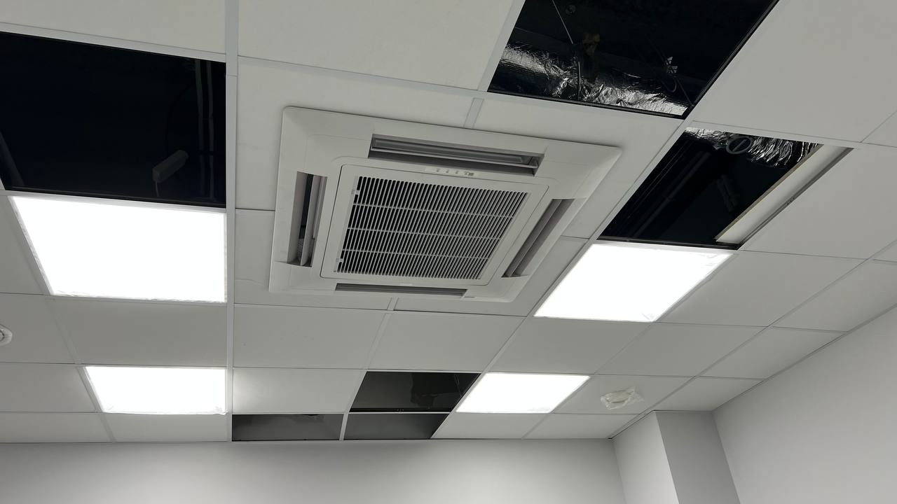 Kasetonowa sufitowa instalacja z centralnie umieszczonym panelem klimatyzacyjnym, otoczonym panelami oświetleniowymi i otworami rewizyjnymi.