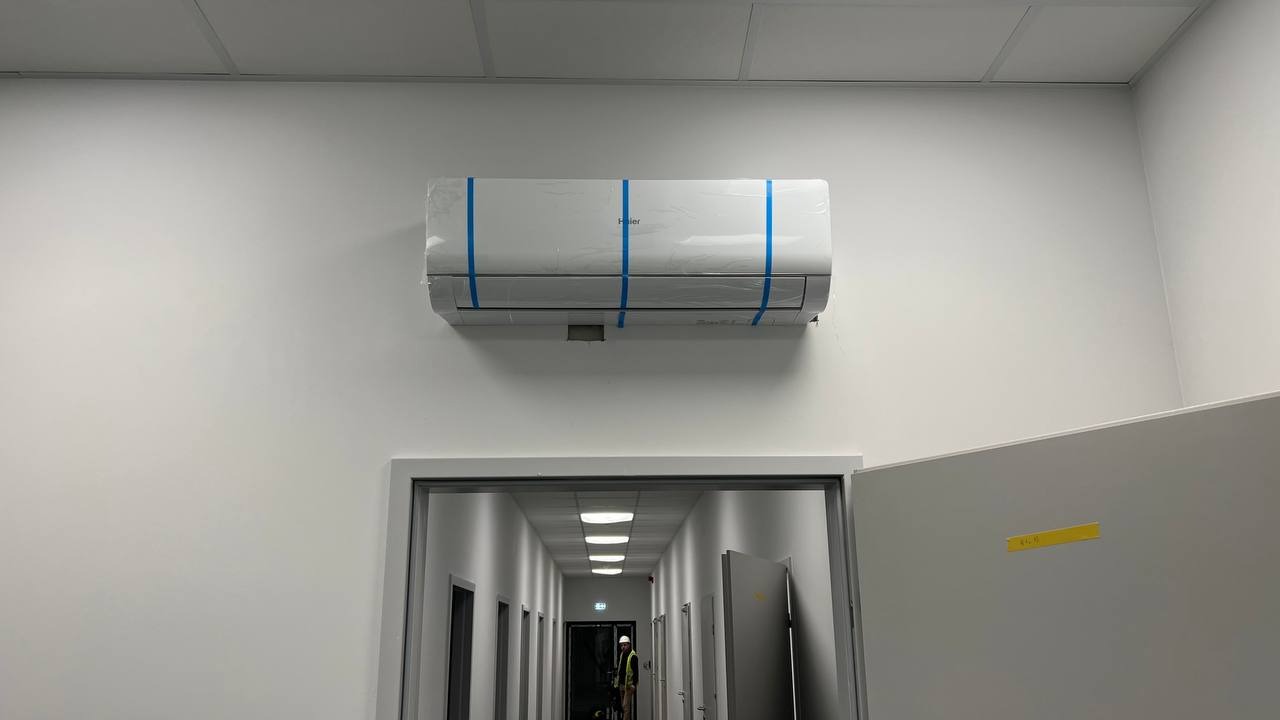 Nowo zamontowana klimatyzacja Haier, jeszcze z folią ochronną i taśmą, w biurowym korytarzu z widoczną osobą w odblaskowej kamizelce w oddali.