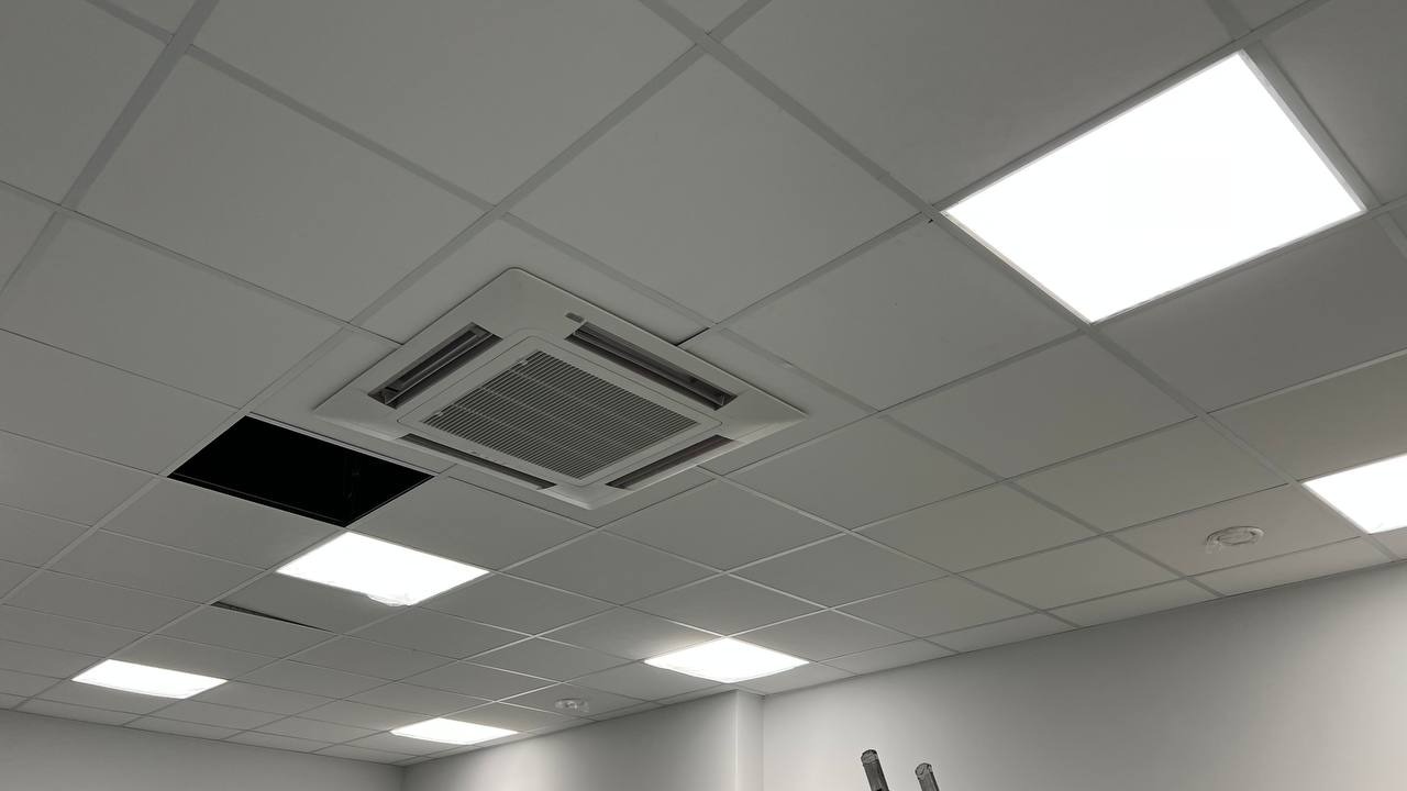 Kasetonowa instalacja sufitowa z wbudowanym klimatyzatorem kasetonowym i panelem oświetleniowym LED; w tle widoczna otwarta przestrzeń w suficie.