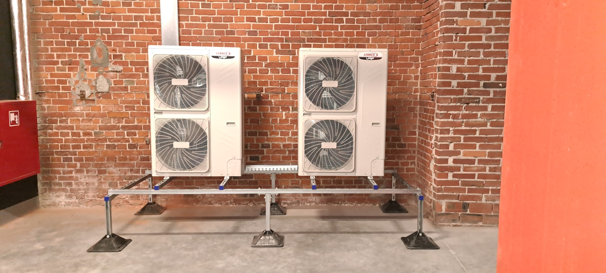 Dwa moduły klimatyzacyjne Lennox VRF ustawione na metalowym stelażu z regulowanymi nóżkami, zamontowane przy ceglanej ścianie w surowym wnętrzu.