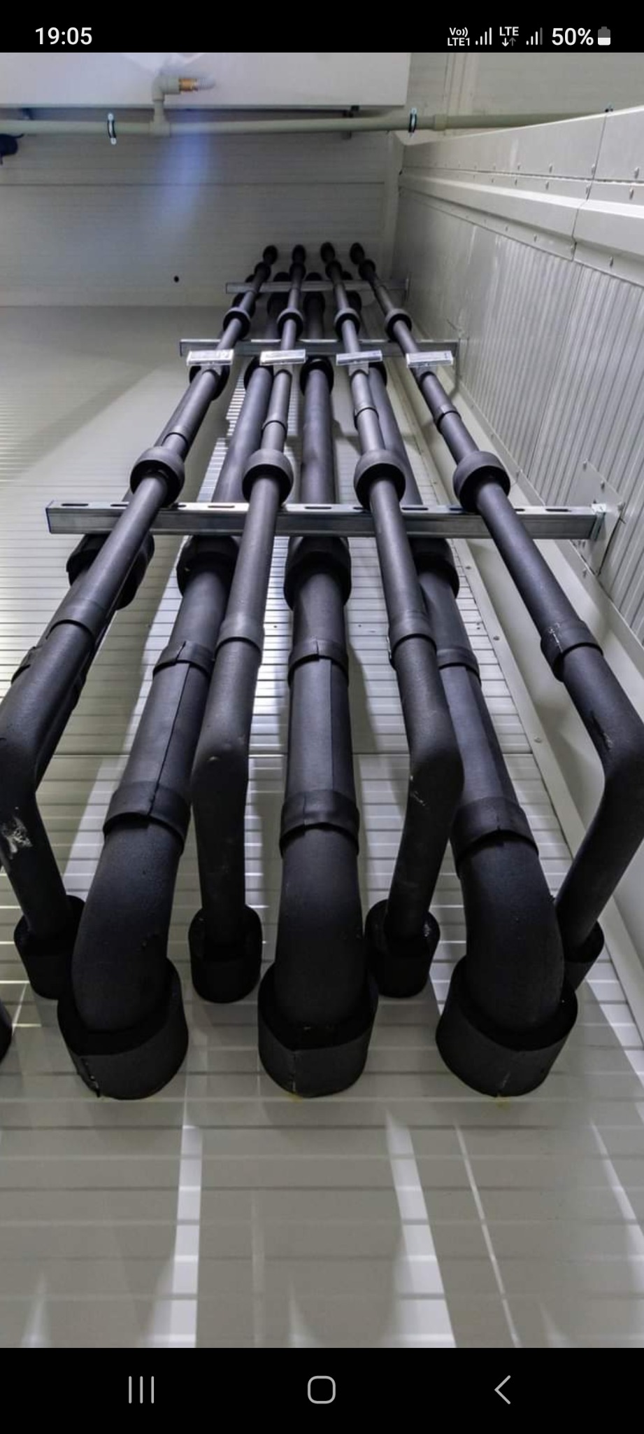 Rzędy zaizolowanych, czarnych rur wentylacyjnych, podwieszonych pod sufitem w pomieszczeniu o białych ścianach z metalowymi panelami, widoczne wsporniki mocujące.