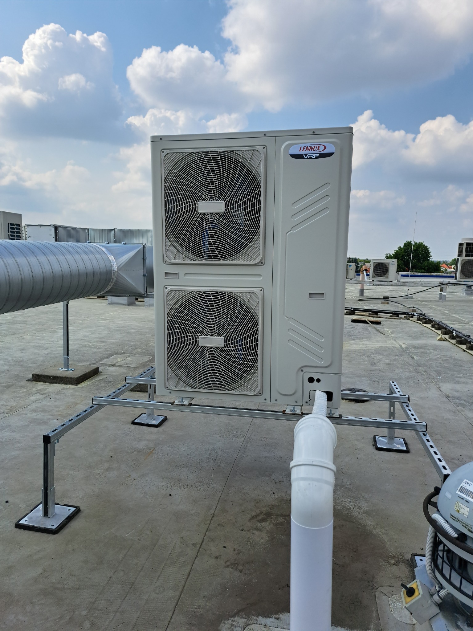 Agregat klimatyzacyjny marki Lennox VRF zamontowany na dachu budynku, widoczny system rur i wsporników, w tle niebo z chmurami.