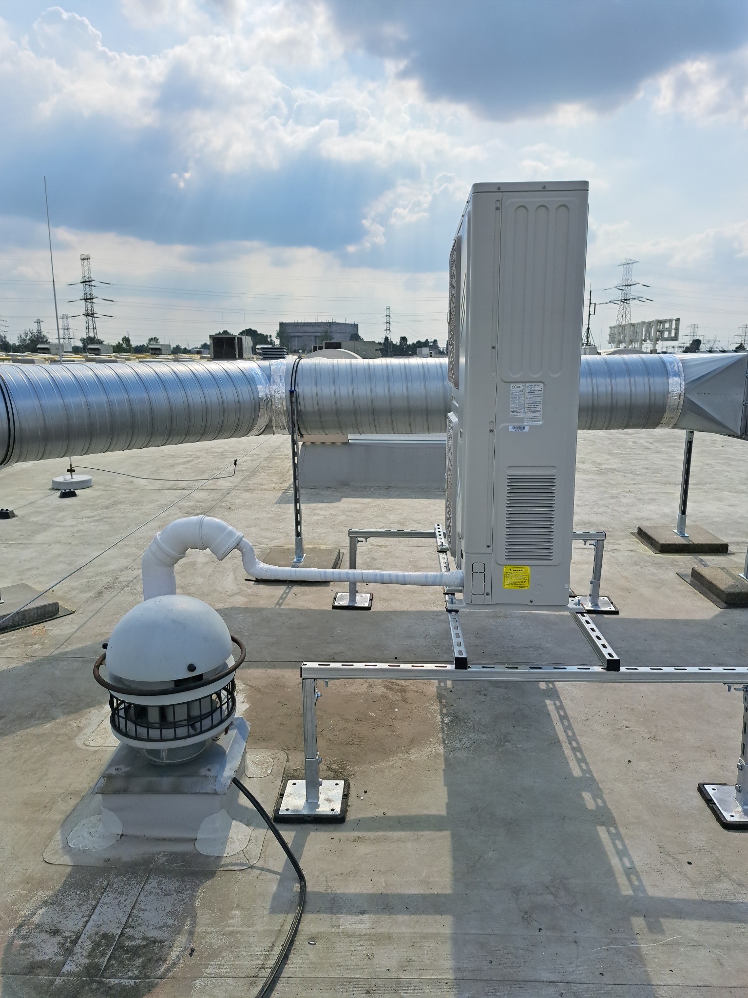 System wentylacyjny na dachu budynku, widoczny centralny moduł, rury wentylacyjne z izolacją oraz element wentylacyjny kulisty z kratką, w tle słupy wysokiego napięcia pod błękitnym niebem.