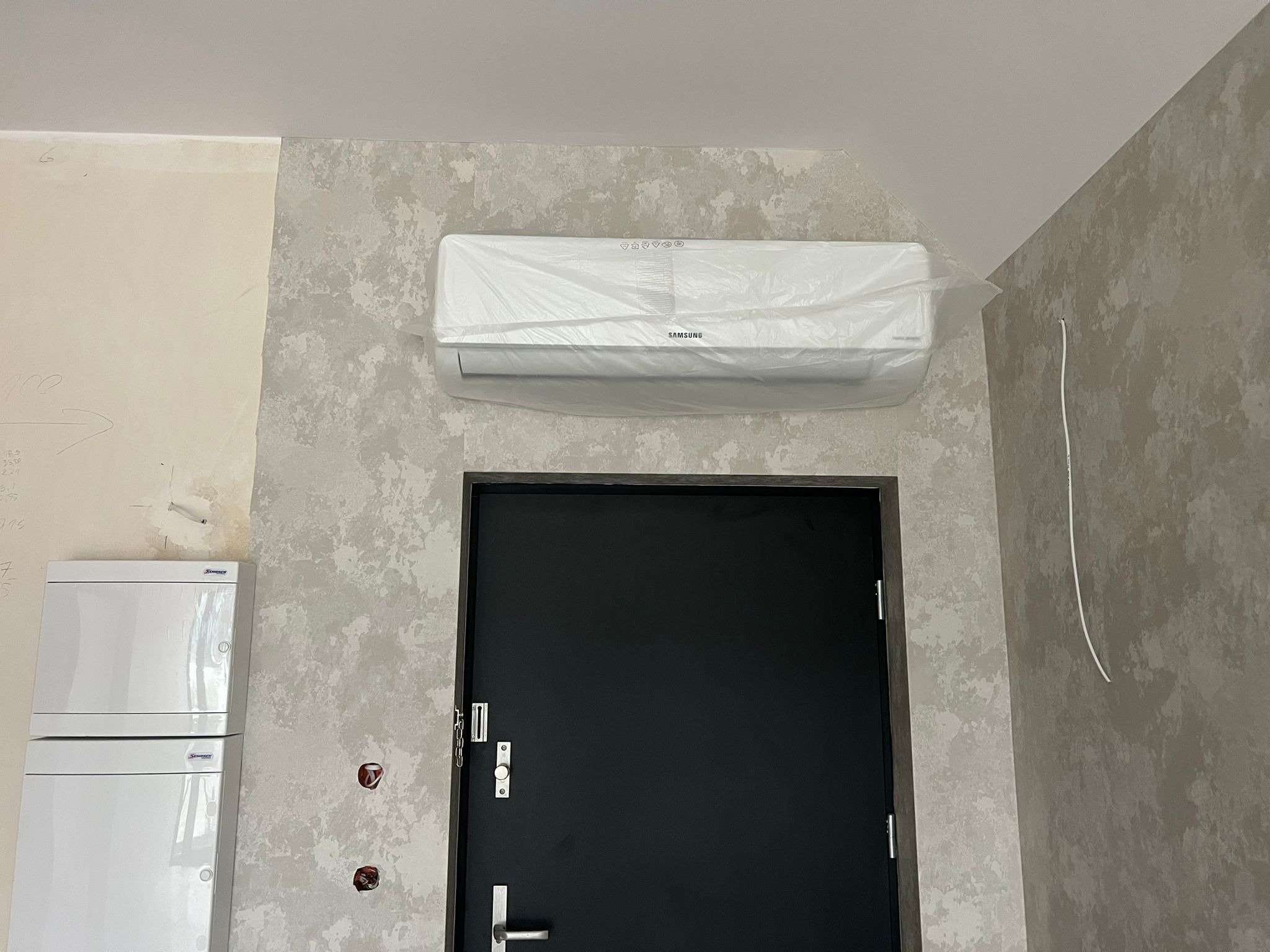 Biała jednostka klimatyzacyjna Samsung, owinięta folią ochronną, zamontowana nad czarnymi drzwiami w pomieszczeniu z dekoracyjną ścianą o szarym wzorze.