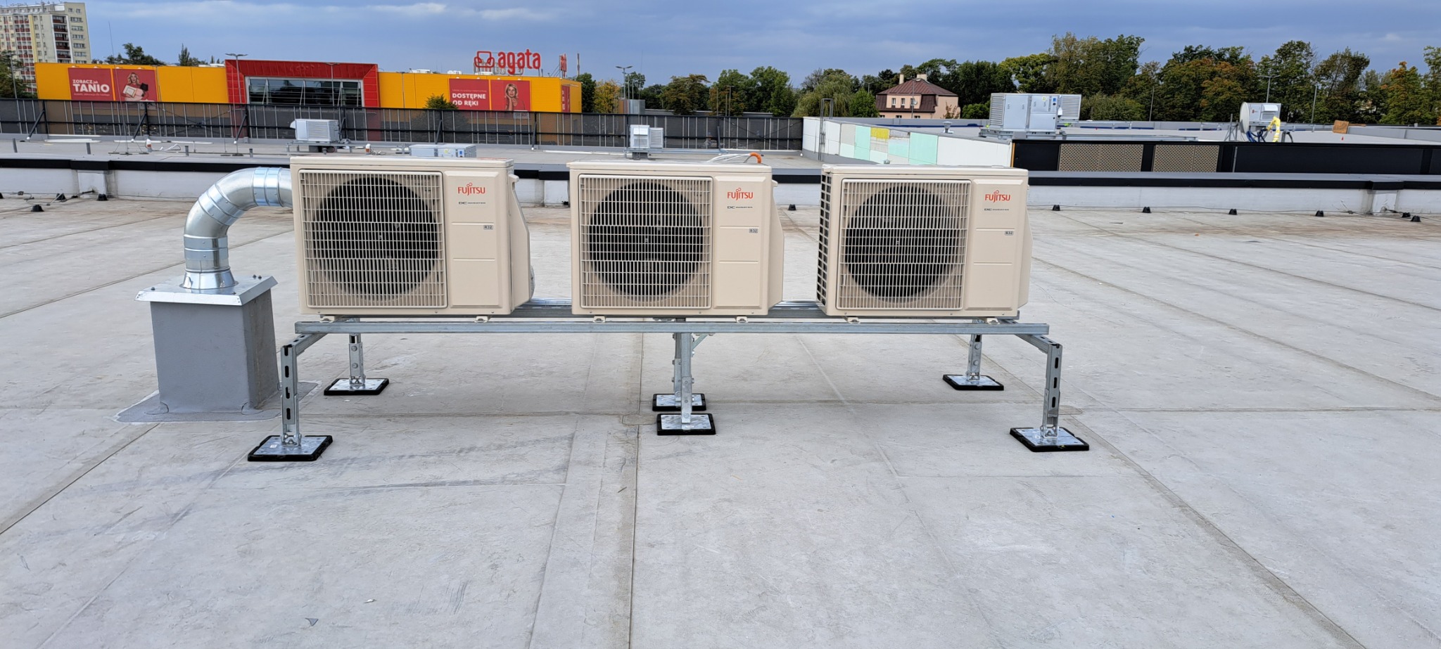 Trzy jednostki zewnętrzne klimatyzacji Fujitsu zamontowane na metalowej konstrukcji na dachu budynku, z widocznym systemem odprowadzania powietrza i budynkami w tle.