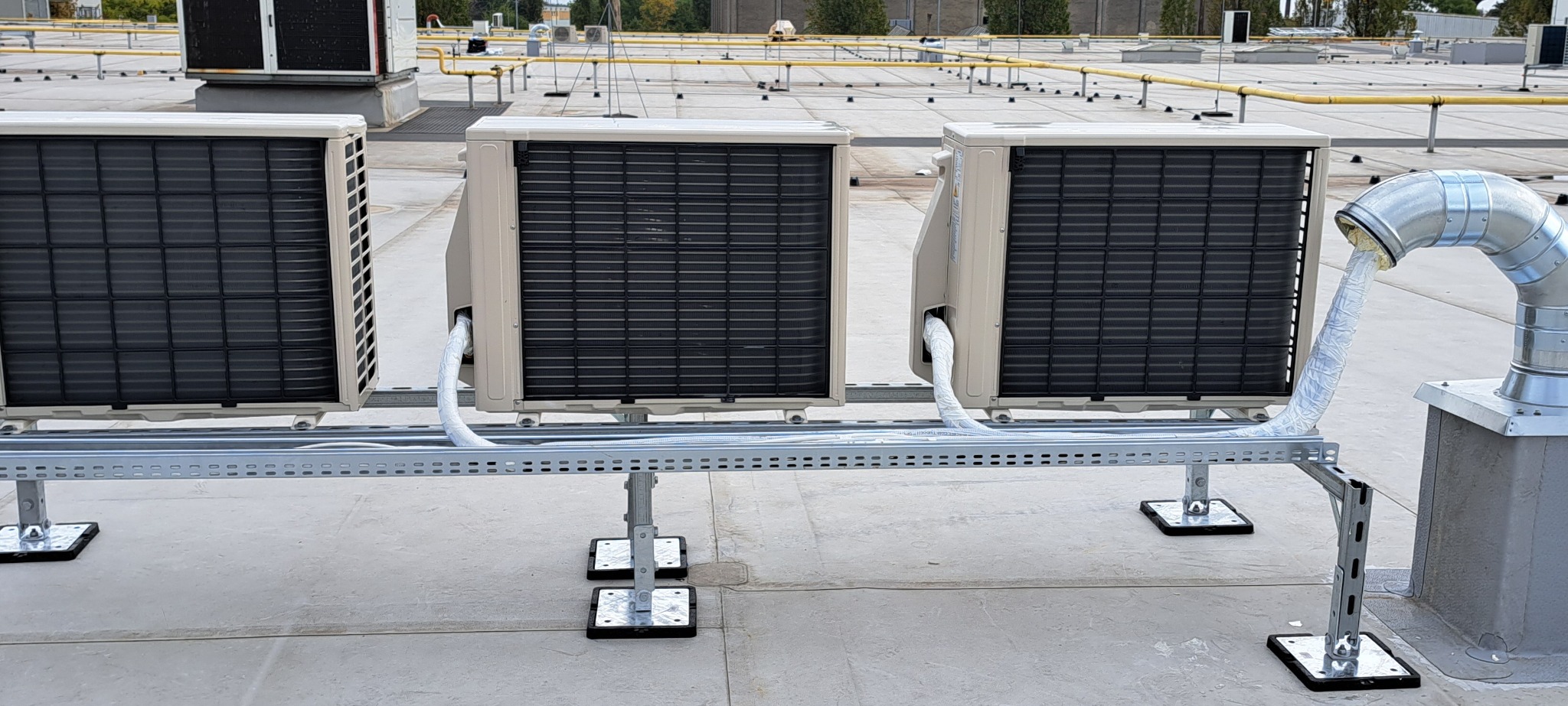 Trzy jednostki zewnętrzne klimatyzacji zamontowane na płaskim dachu, połączone rurami w izolacji, widoczny system wsporników i mocowań.