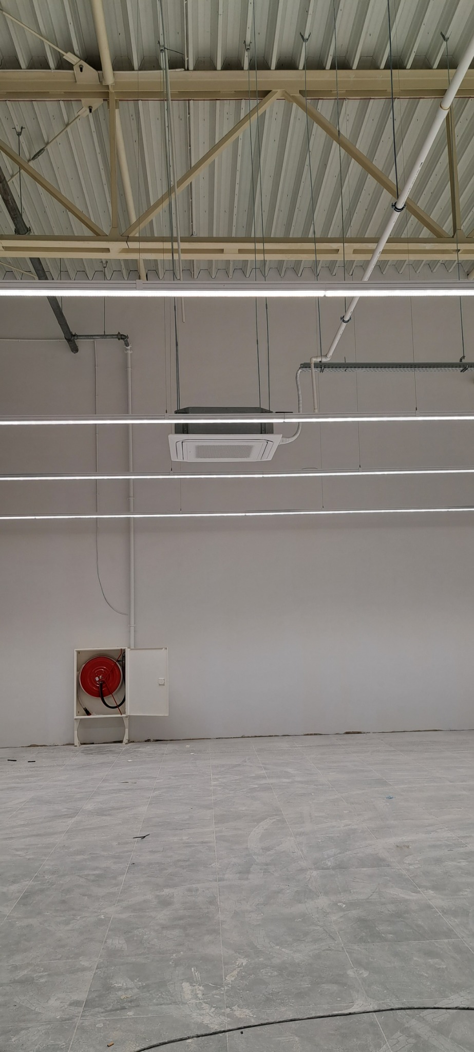 Sufit w hali przemysłowej z zamontowaną kasetą klimatyzacyjną, oświetleniem ledowym, instalacją przeciwpożarową z wężem w szafce i szarą podłogą z płytek.