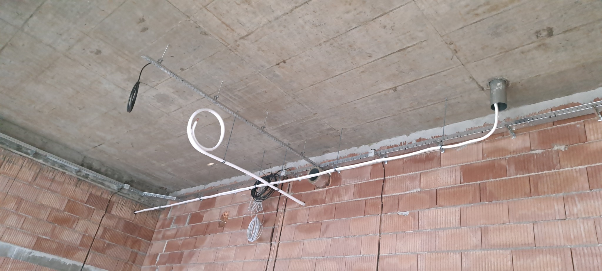 Montaż instalacji wentylacyjnej: białe rury prowadzone pod betonowym sufitem, mocowane do metalowych szyn na tle ściany z czerwonej cegły.