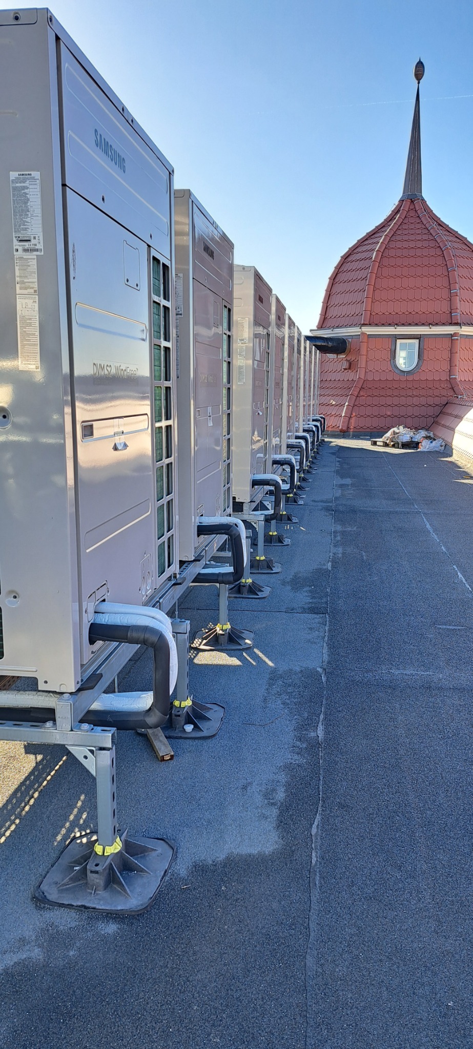 Rząd zewnętrznych jednostek klimatyzacyjnych Samsung DVM S2 WindFree ustawionych na dachu budynku z czerwoną wieżyczką w tle.