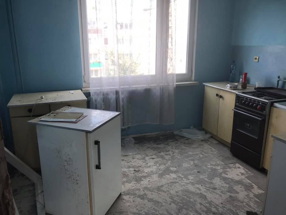 Kuchenne wnętrze przed renowacją: niebieskie ściany, stare szafki, odsłonięta podłoga z pozostałościami kleju, okno z firanką.