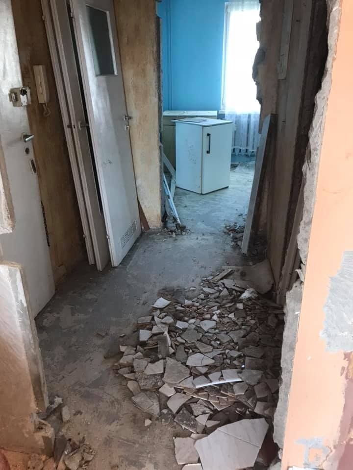 Korytarz w trakcie demontażu, widoczne fragmenty gruzu, drzwi i ściany w stanie surowym, w tle lodówka.