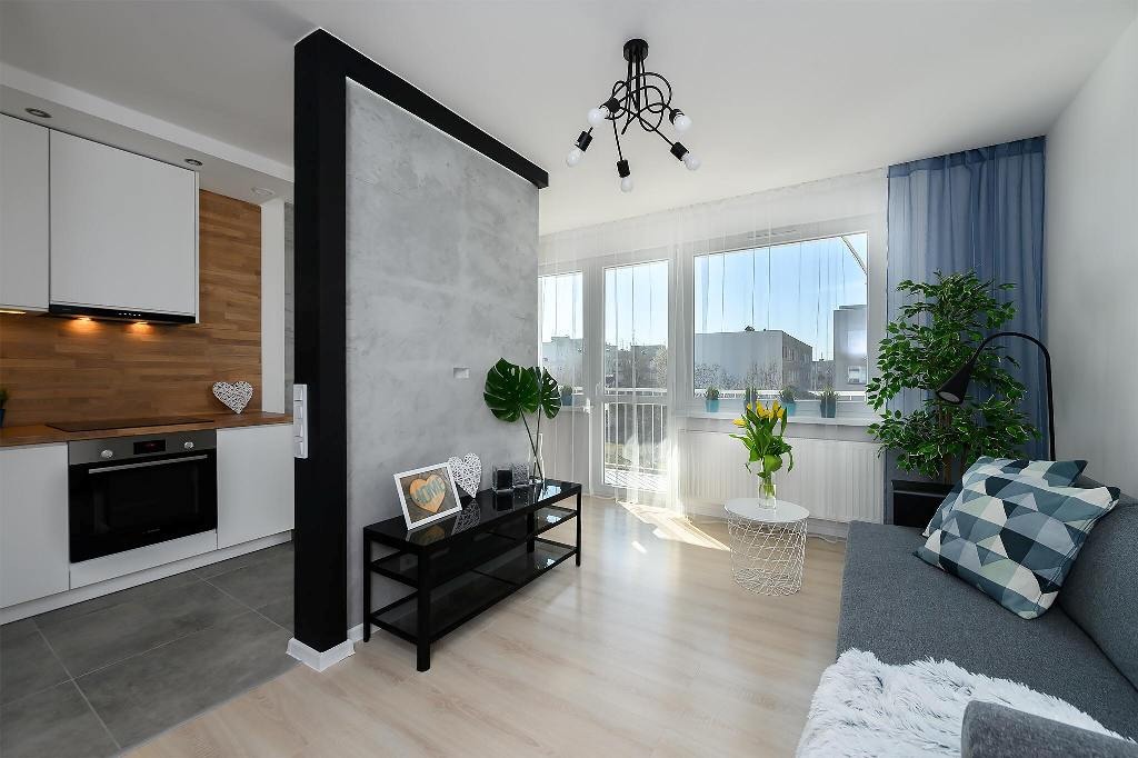 Jasne wnętrze mieszkania z szarą ścianą dekoracyjną, drewnianym panelem w kuchni i nowoczesnym oświetleniem, widok z perspektywy salonu na kuchnię.