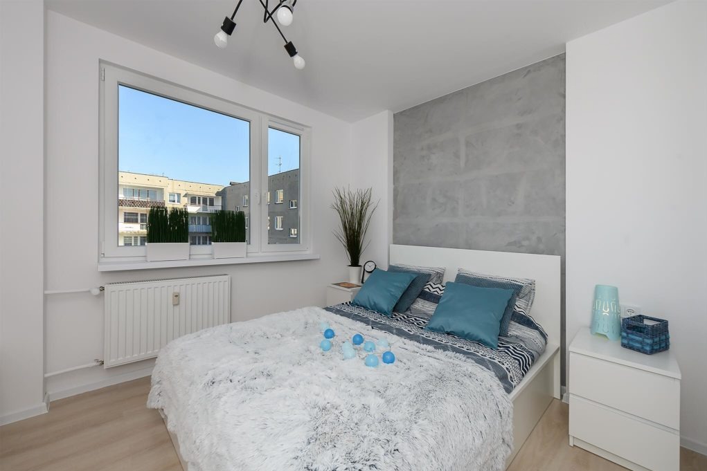 Nowoczesna sypialnia z łóżkiem, dekoracyjną ścianą imitującą beton, białymi meblami i widokiem na budynek mieszkalny z zielenią na balkonie.