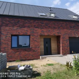 Nowoczesny dom jednorodzinny z czerwoną ceglaną elewacją, grafitowym dachem z lukarnami, drewnianym wykończeniem wejścia, szarą bramą garażową i stosem kostki brukowej przed budynkiem.