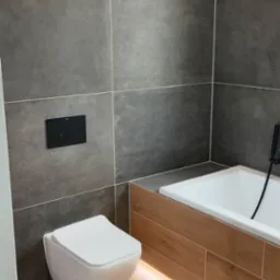 Nowoczesna łazienka z szarymi, matowymi płytkami na ścianach, podwieszaną toaletą i podświetlaną wanną obudowaną płytkami imitującymi drewno.