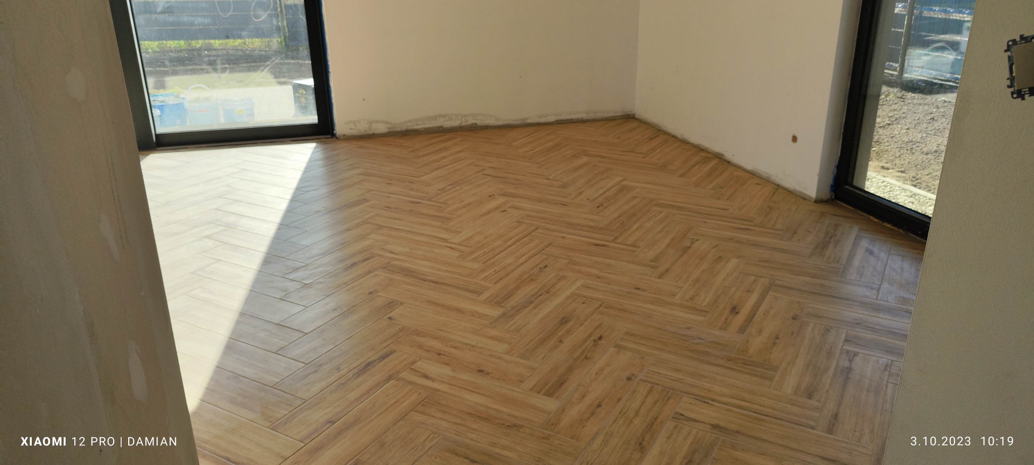 Ułożona podłoga z płytek imitujących drewno w jodełkę, widoczne niedoskonałości wykończenia ścian i narożników, przygotowanie do dalszych prac wykończeniowych.