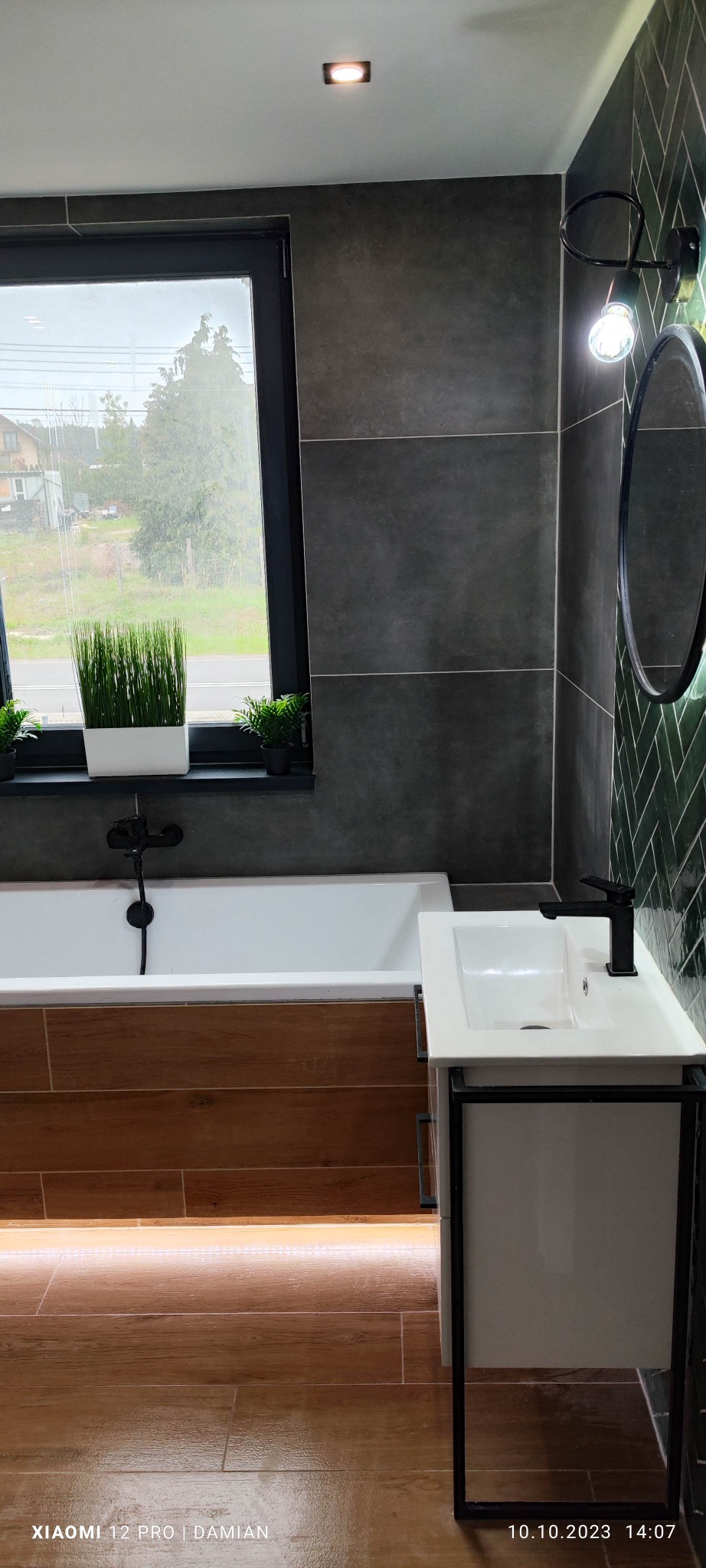 Nowoczesna łazienka po remoncie: biała wanna z drewnianą obudową, umywalka nablatowa z czarną armaturą, szara ściana z dużych płytek, dekoracyjne rośliny w oknie i podświetlana podłoga z płytek...