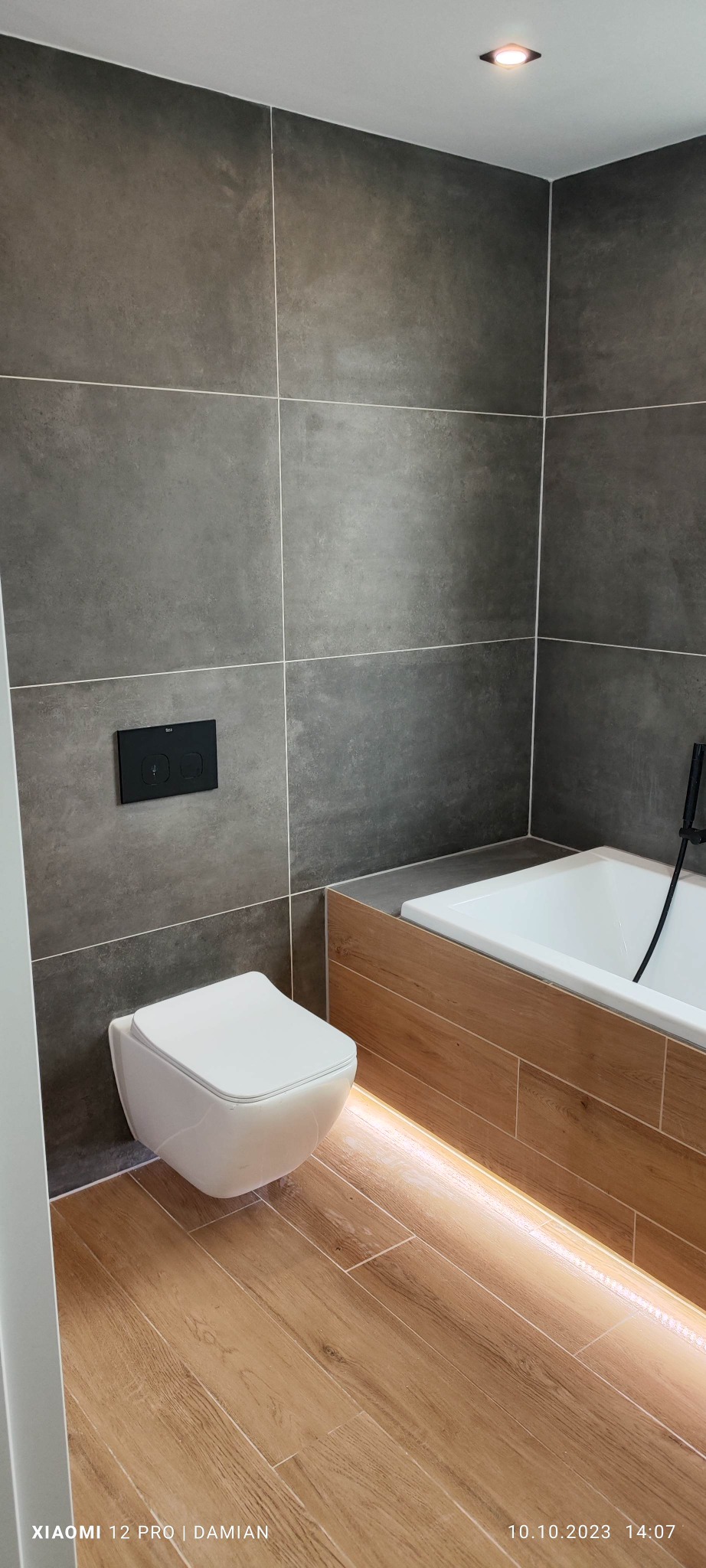 Nowoczesna łazienka z szarymi, matowymi płytkami na ścianach, podwieszaną toaletą i podświetlaną wanną obudowaną płytkami imitującymi drewno.