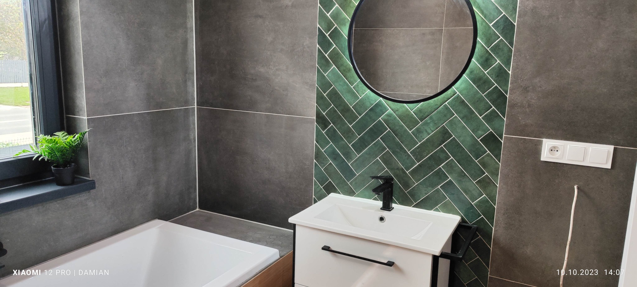 Nowoczesna łazienka z szarymi płytkami wielkoformatowymi, zieloną ścianą z płytek ułożonych w jodełkę, okrągłym lustrem z podświetleniem LED, białą szafką z umywalką i czarną armaturą oraz rośliną...