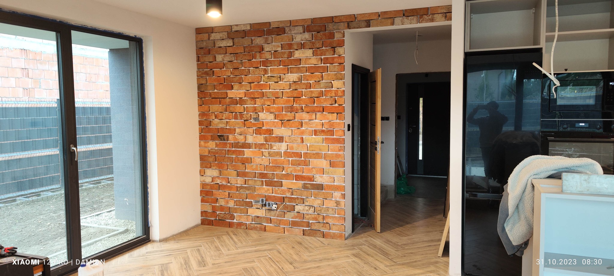 Wnętrze w trakcie wykańczania: ściana z cegły klinkierowej, parkiet ułożony w jodełkę, widoczne okno z zewnątrz oraz częściowo zabudowana kuchnia z czarnym frontem.