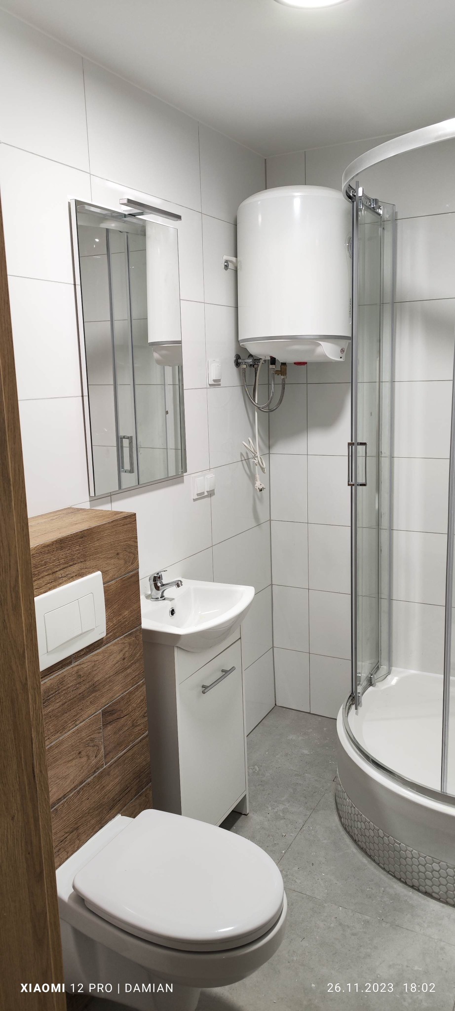 Nowoczesna łazienka z białą armaturą, kabiną prysznicową z zaokrąglonymi drzwiami, podgrzewaczem wody Atlantic i płytkami w jasnych odcieniach szarości i bieli.