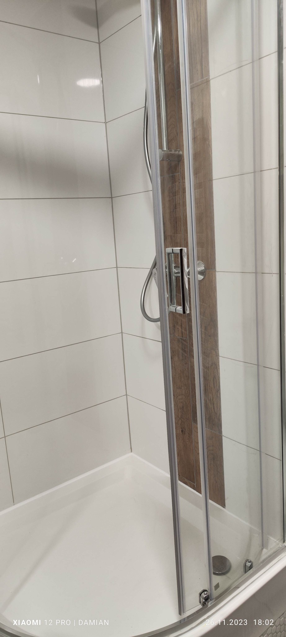 Kabina prysznicowa z przesuwnymi drzwiami, wykończona płytkami w kolorze białym i panelem imitującym drewno, widoczny chromowany uchwyt i bateria.