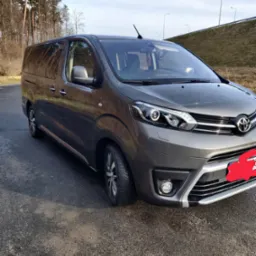 Szary van Toyota Proace Verso zaparkowany na asfalcie, widok z przodu, w tle drzewa i nasyp z barierami drogowymi.