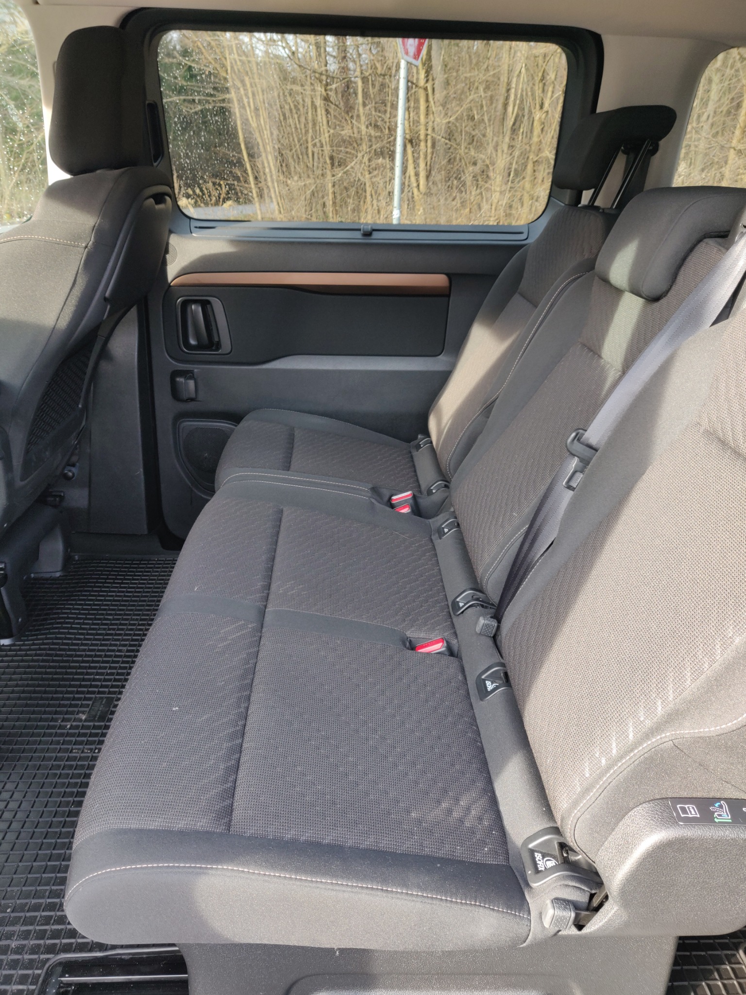 Wnętrze samochodu osobowego z trzema rzędami siedzeń, widoczne czarne, gumowe dywaniki oraz zagłówki i pasy bezpieczeństwa, za oknem drzewa bez liści.