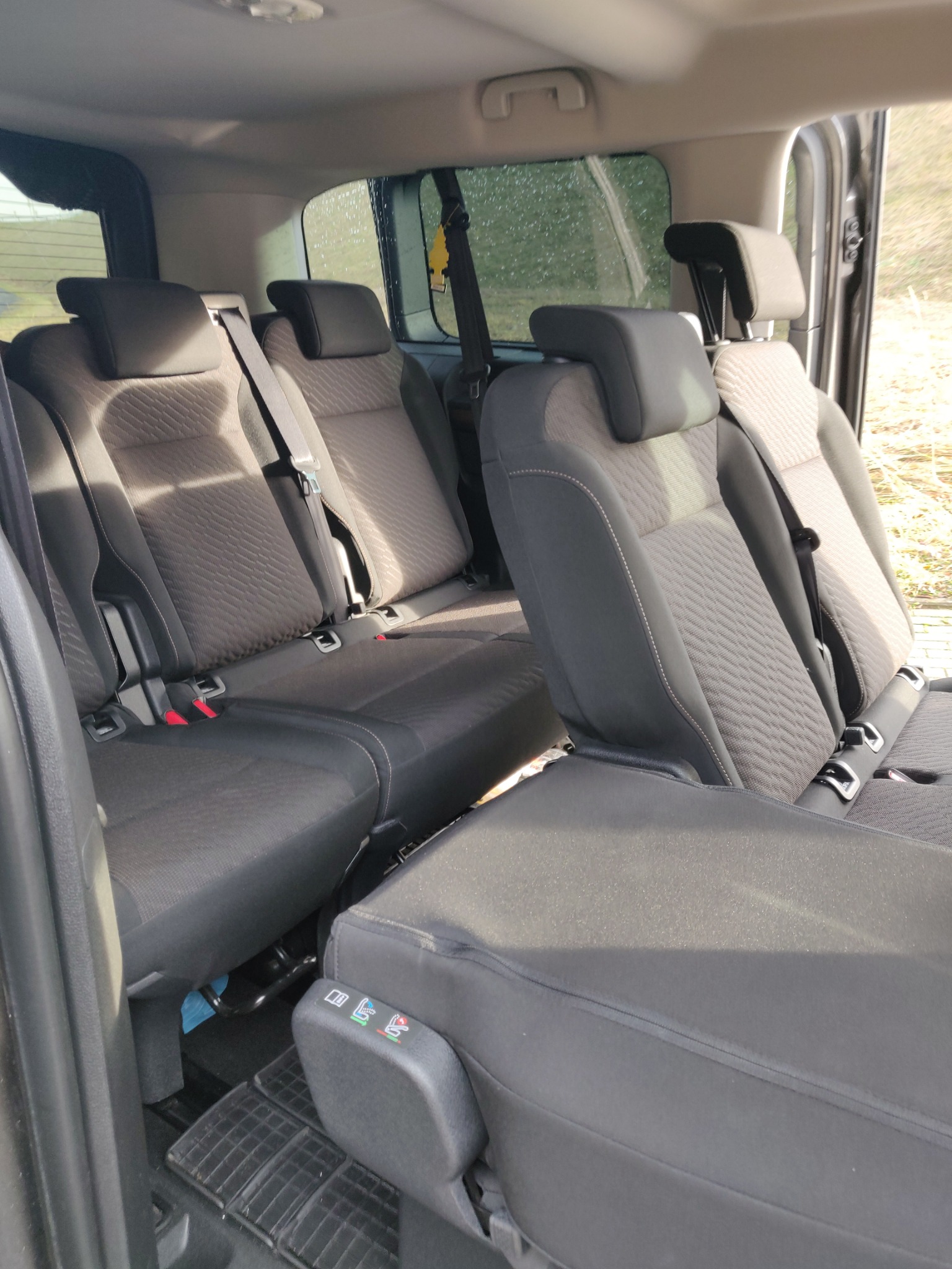 Wnętrze samochodu typu van z trzema rzędami siedzeń w kolorze ciemnoszarym, z widocznymi zagłówkami, pasami bezpieczeństwa i elementami regulacji foteli.