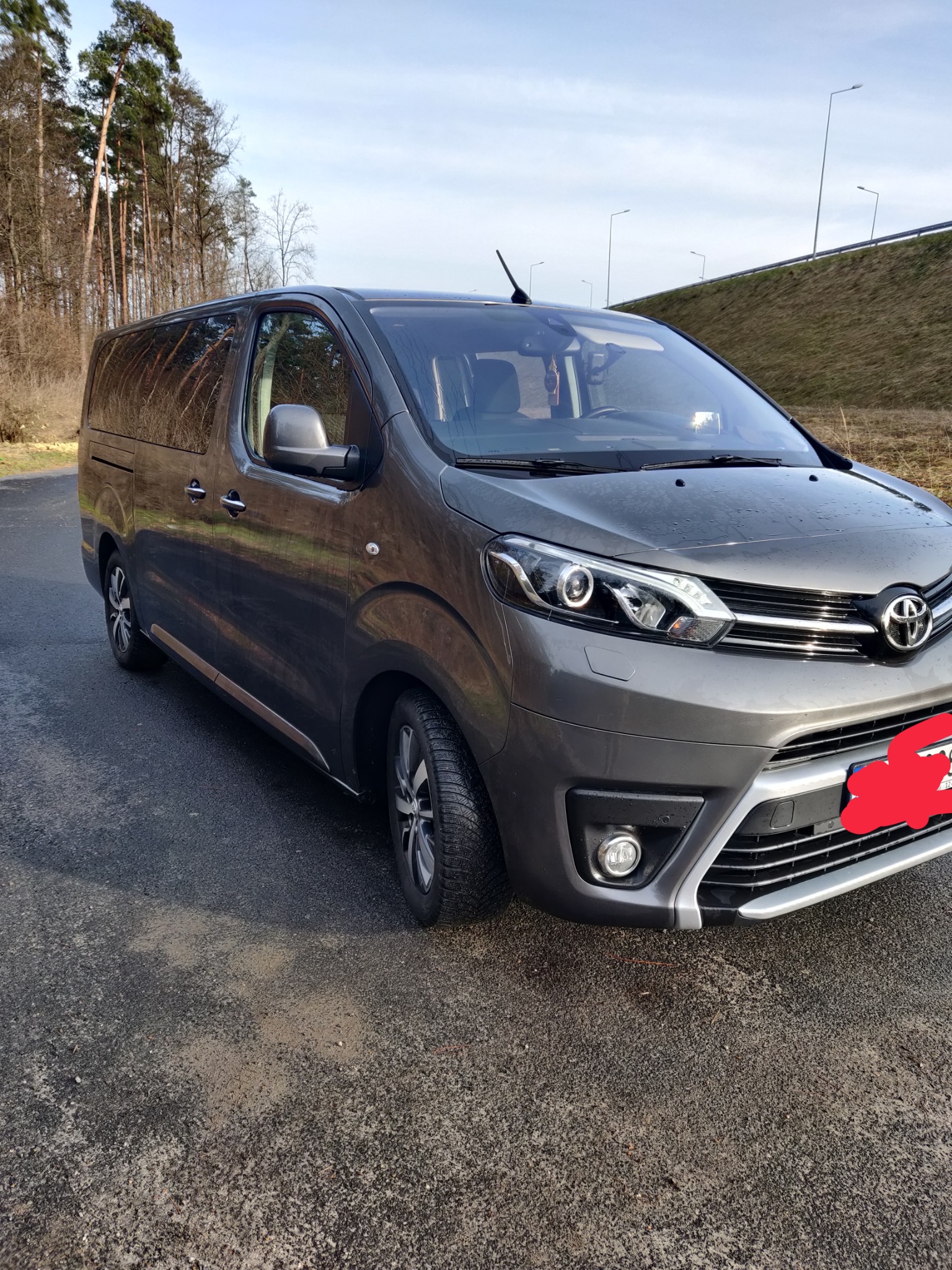 Szary van Toyota Proace Verso zaparkowany na asfalcie, widok z przodu, w tle drzewa i nasyp z barierami drogowymi.